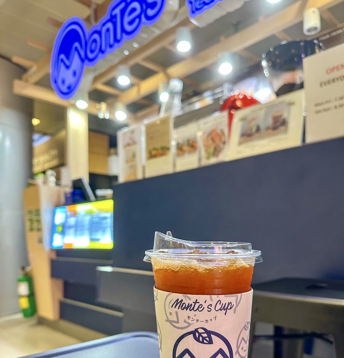 เคาน์เตอร์ Monte’s Cup ที่สนามบิน มีป้ายไฟนีออนโลโก้ร้าน และแก้วเครื่องดื่มที่สวมปลอกแก้วลายแบรนด์